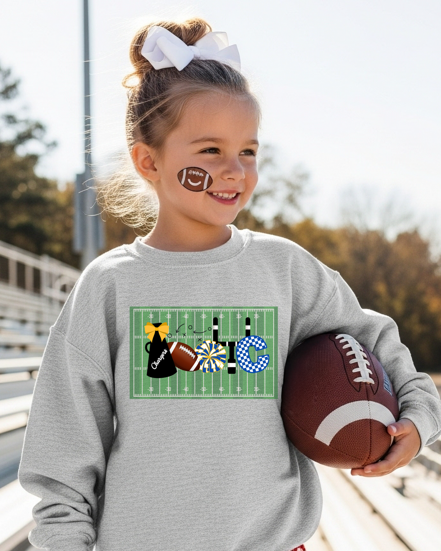 Young girl holding a football and wearing a sweatshirt with a sports-themed design.
