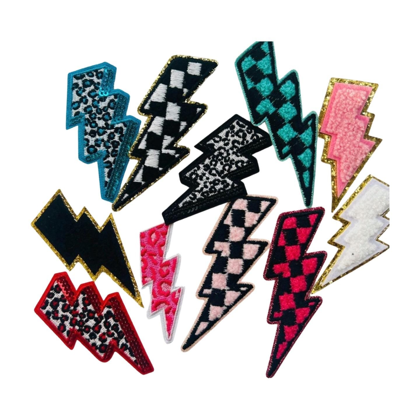 May include: A collection of 12 lightning bolt shaped iron on patches in various colors and patterns including pink, black, white, blue, and red. The patches have checkered, leopard print, and fuzzy textures.