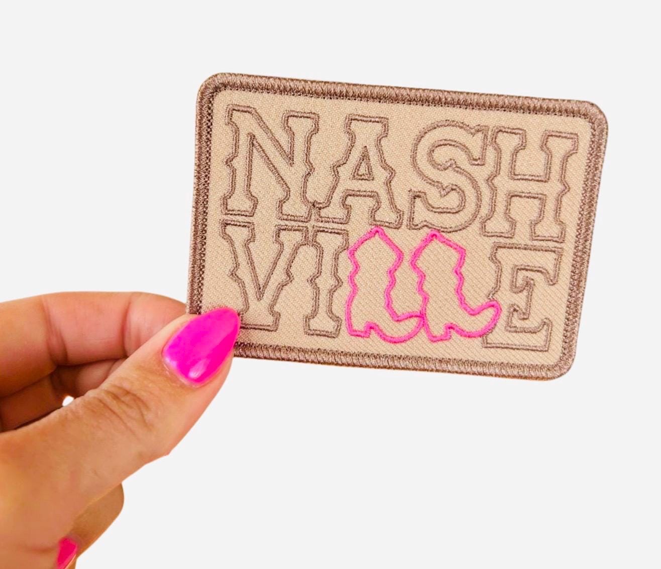 May include: A tan fabric patch with the word &#39;NASHVILLE&#39; embroidered in brown thread. The word &#39;VILLE&#39; is outlined in pink thread and has a pair of pink cowboy boots embroidered in the center.