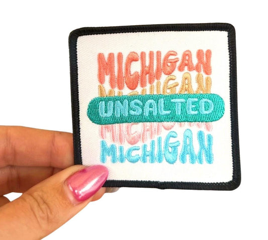 May include: A white square patch with black border. The patch has the word &#39;MICHIGAN&#39; written in pink, orange, and yellow. The word &#39;UNSALTED&#39; is written in green. The word &#39;MICHIGAN&#39; is written in blue.