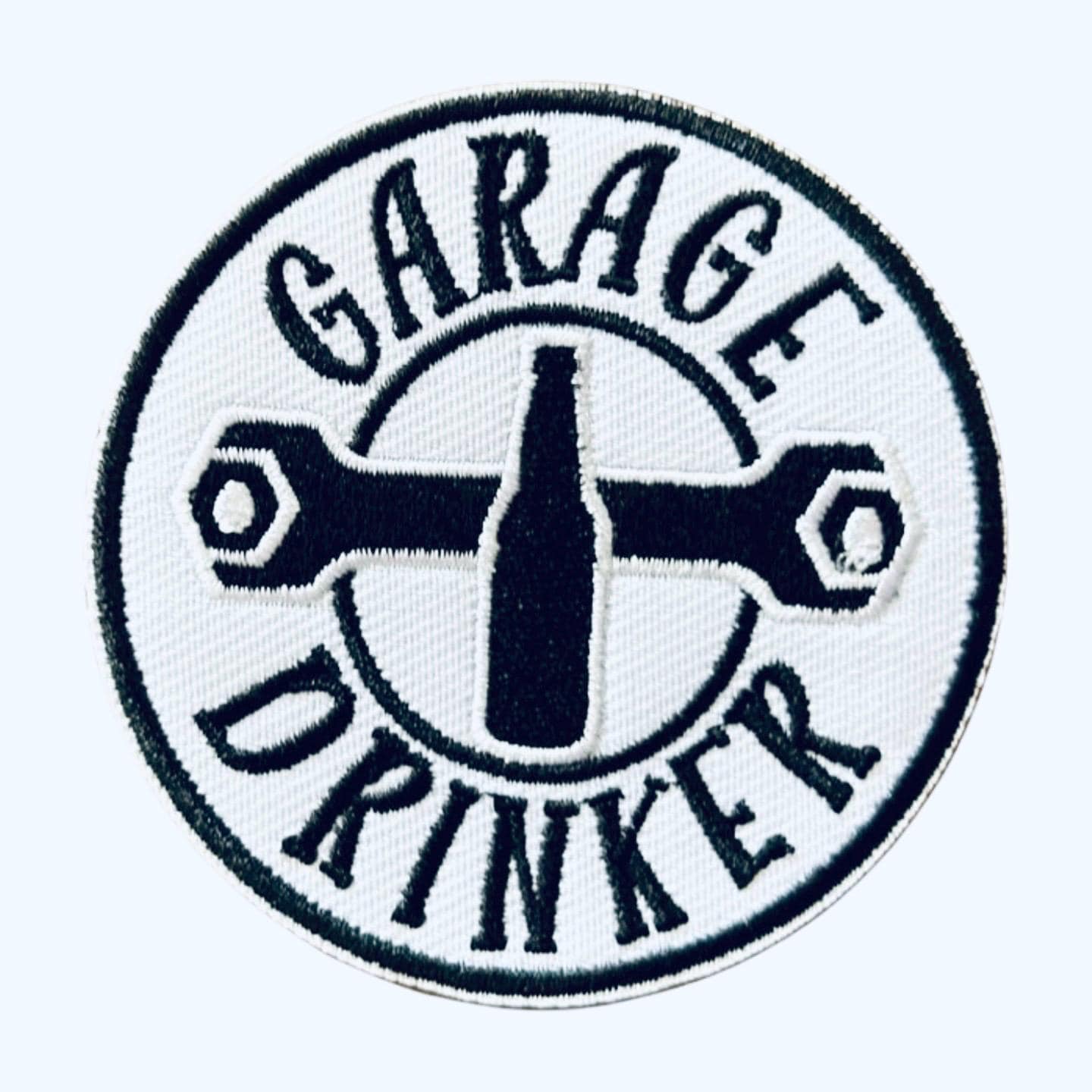 May include: A white circular patch with black embroidered text and a wrench design. The text reads &#39;Garage Drinker&#39; with a bottle shape in the center.