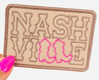 May include: A tan fabric patch with the word &#39;NASHVILLE&#39; embroidered in brown thread. The word &#39;VILLE&#39; is outlined in pink thread and has a pair of pink cowboy boots embroidered in the center.