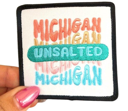 May include: A white square patch with black border. The patch has the word &#39;MICHIGAN&#39; written in pink, orange, and yellow. The word &#39;UNSALTED&#39; is written in green. The word &#39;MICHIGAN&#39; is written in blue.