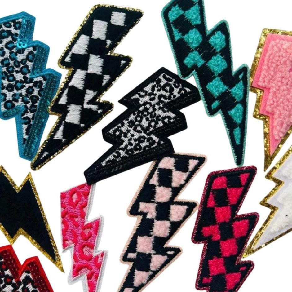 May include: A collection of 12 lightning bolt shaped iron on patches in various colors and patterns including pink, black, white, blue, and red. The patches have checkered, leopard print, and fuzzy textures.