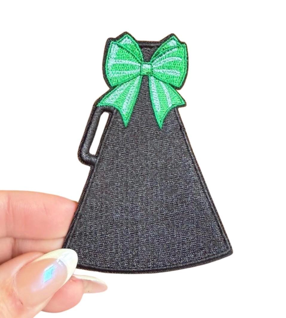 Black dress-shaped patch with a green bow held by a hand on a white background