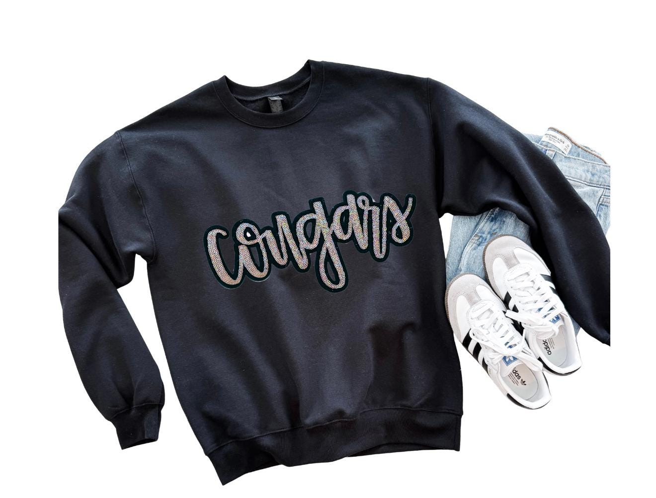 Black sweatshirt with 'Cougars' text, white sneakers, and jeans on a white background